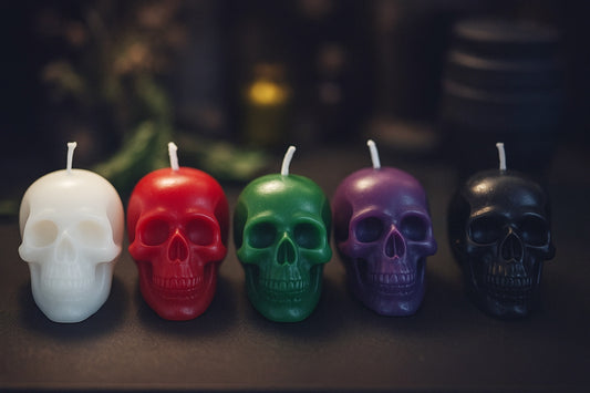 Five skull-shaped ritual candles in white, red, green, purple, and black arranged in a row on a dark altar surface, moody lighting highlighting their glossy carved details for gothic ritual decor.